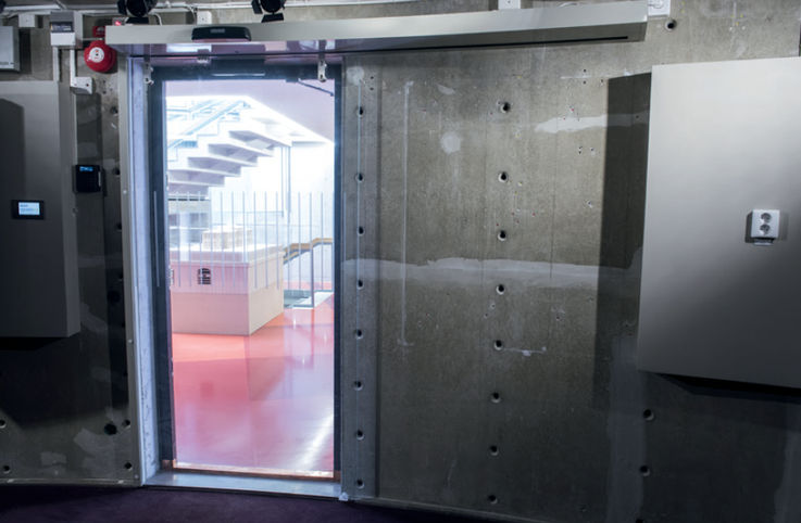 Une porte coulissante en verre commandée par un capteur dans un mur en béton donnant sur une zone d'escalier lumineuse.