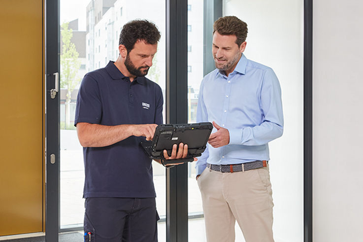 In a modern entrance area, a service employee shows a customer in business clothing technical information on a laptop. 
